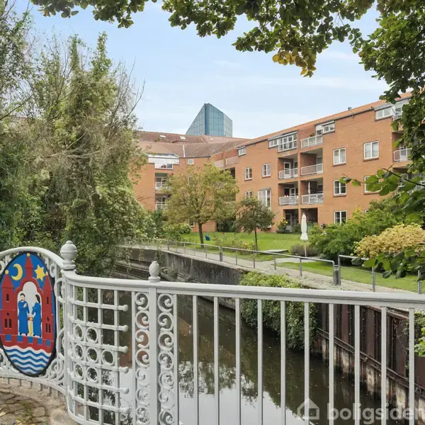 Et dekorativt rækværk med et farverigt maleri står ved en lille flod. I baggrunden ses murstensbygninger og grønne træer under en klar himmel.