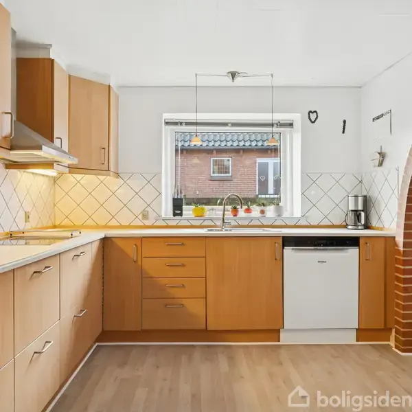 Et lyst køkken med træskabe og opvaskemaskine, der står under et vindue med udsigt til en bygning. Et murstensbuet arkade fører til et tilstødende rum.