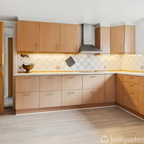 Køkkenskabe i lyst træ med indbygget ovn og emhætte på væggen, omgivet af hvide fliser og en frugtskål på bordet. Opvaskemaskine ved siden af et stort vindue.