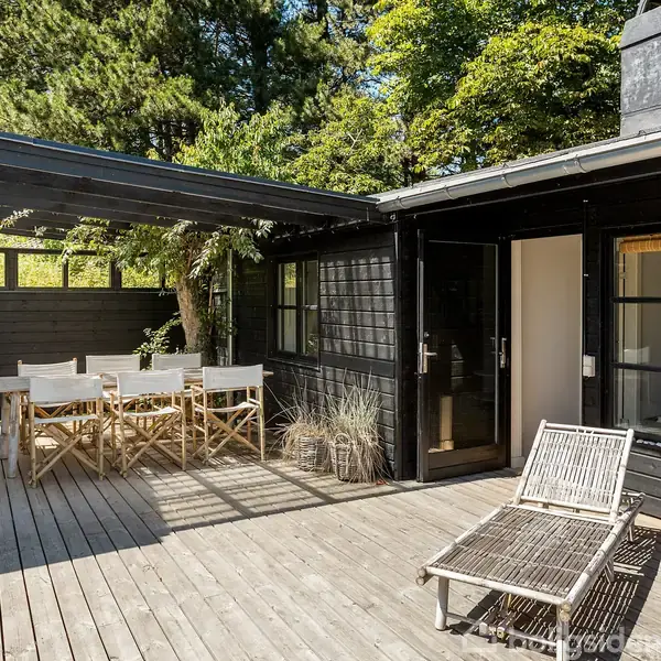 En træterrasse med sortmalet hus, hvorpå der står et bord og seks stole under et halvtag. En liggestol er placeret ved en åbnet dør. Omgivet af træer.