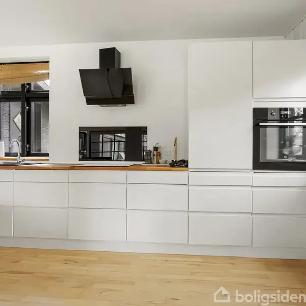 Et moderne køkken har hvide skabe og træbordplade. Der er sort emhætte, mikroovn og ovn indbygget. Vinduer med persienner giver naturligt lys. Hårde hvidevarer står på bordet.