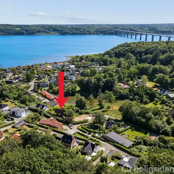 Rødt hus vises med en rød pil pegende mod det. Det ligger i et grønt, beboeligt område nær en bred flod med en bro i baggrunden.