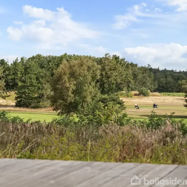Stol på terrasse overlooket af frodig natur, mens to personer cykler på en sti i et skovklædt landskab under en klar blå himmel.