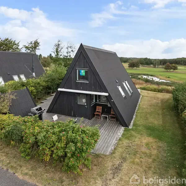 Sort A-formet hus med træbeklædning står på en plæne, omgivet af buske. En terrasse med havemøbler ses foran, og i baggrunden er der grønne marker og træer under en blå himmel.