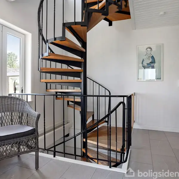 En sort, moderne vindeltrappe fører opad i en lys gang med grå flisegulv. En kurvestol står ved vinduet, og en plakat hænger på væggen.