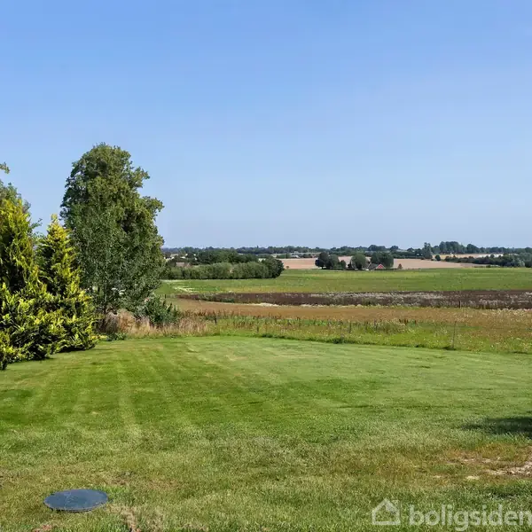 Græsplæne strækker sig ud mod horisonten, omgivet af marker og et par træer til venstre, under en klar blå himmel.