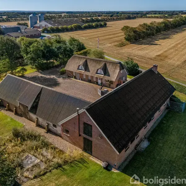 Stort, traditionelt landhus med stråtækt tag omgivet af landbrugsbygninger. Bygningerne er beliggende i et åbent landskab med marker, træer og en gårdsplads.