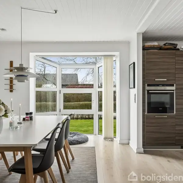 Spisebord med sorte stole står i en lys og moderne spisestue, hvor to døre åbner ud mod en grøn have. Køleskab og ovn er indbygget i køkkenskabene.