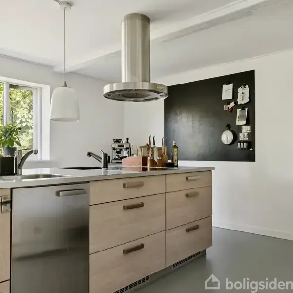 Et gråt hus med en skrånende tagflade har to vinduer. Foran huset er der en grusbelagt gårdsplads og nogle planter. Omgivelserne inkluderer træer og grønt løv.