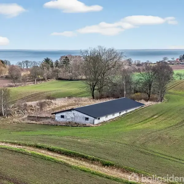 En lang, hvid bygning med sort tag står stille på en grøn bakke i et bakket landskab. I baggrunden ses en skov og havet under en lys, blå himmel.