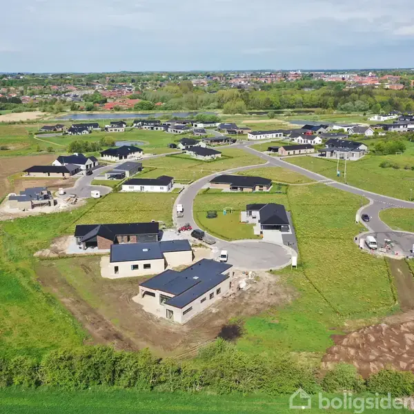 Flere moderne enfamiliehuse under opførelse ligger i et åbent, grønt forstadsområde. En vej snor sig mellem dem, omgivet af græsarealer og marker, med en by i baggrunden.