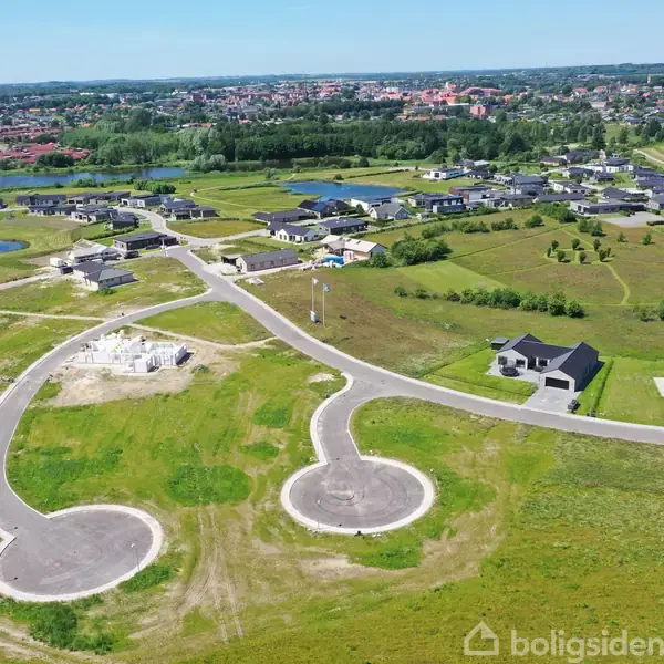 To rundkørsler er forbundet med en vej i et nyt boligområde under opbygning, omgivet af grønne marker og spredte bygninger. I baggrunden ses en by med flere huse og træer.