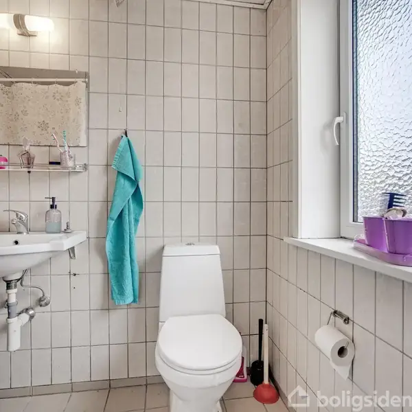 Toilet står på et flisebelagt badeværelse med en håndvask og spejl ved siden af. Et turkis håndklæde hænger, og en skiftelighed af toiletartikler står på vindueskarmen.