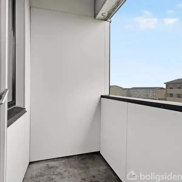 Balkon med hvidt rækværk og betongulv. Den er delvist indrammet af en bygning. I baggrunden ses andre boligblokke mod en lys, skyfri himmel.