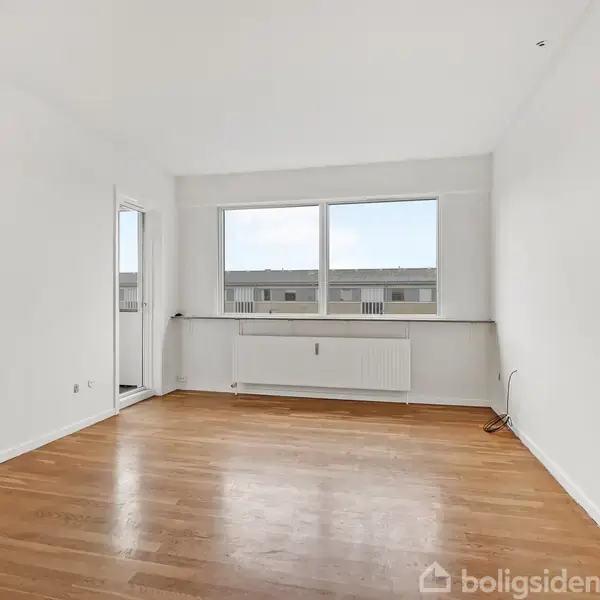 Et tomt rum med trægulv og hvide vægge, indeholder et stort vindue og en radiator. Udsigt til bygninger udenfor. En glasdør til venstre fører til en balkon.
