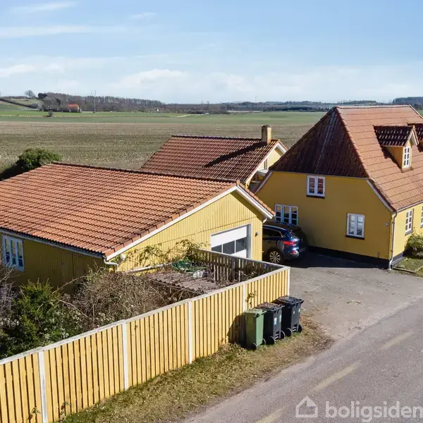 Gulmalet hus med rødt tegltag står stille ved en vej, omkranset af træhegn, med grønne marker i baggrunden. Fire skraldespande står langs indkørslen.