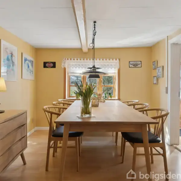 Et langt træbord med stole omkring står i en spisestue. Bordet har en vase med blomster, og en lampe hænger over det. Væggene er gule, og der hænger kunst.