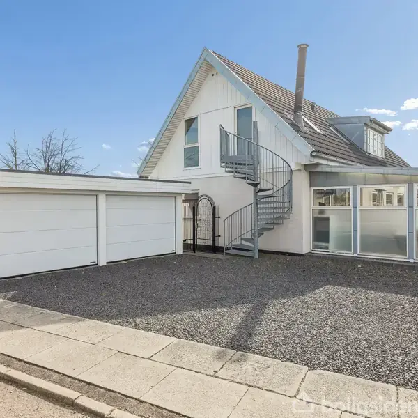 Hvidt hus med en udendørs spiraltrappe, står ved siden af en garage. Gruset indkørsel omkranser huset, mens blå himmel og bare træer ses i baggrunden.