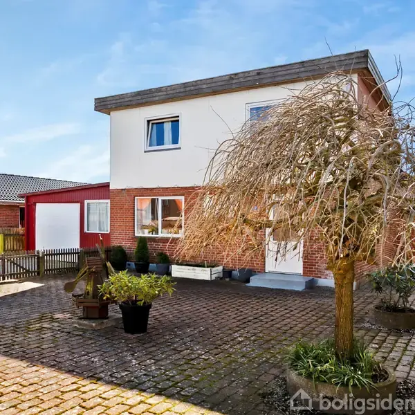 Huset står på en brolagt gårdsplads med flere krukker og et lille træ. Bygningen har to etager, rød murstensfacade og hvide vinduer. En garage og hegn omkranser pladsen.
