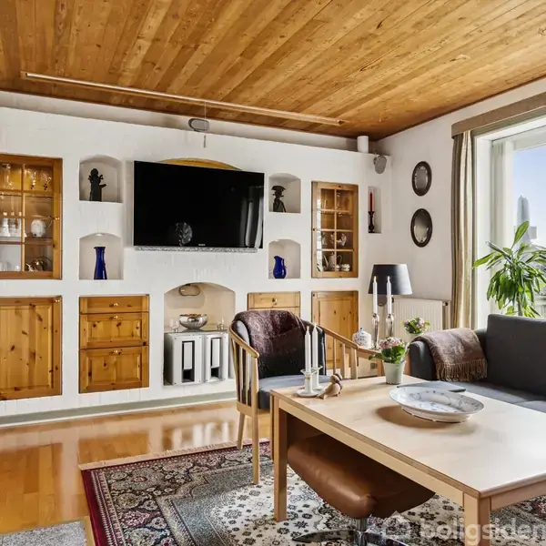 Et moderne stueområde har en sort sofa ved et træbord med tallerkener og glas. En stor fladskærms-TV hænger på væggen med indbyggede skabe. Vindue med udsigt og lyse gardiner.