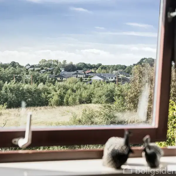 Et åbent vindue vender mod en frodig, grøn udsigt med træer og en række huse i horisonten. To pyntegenstande ses på vindueskarmen.