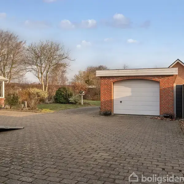 Garage med rød murstensfacade og hvid port står midt i en stort flisebelagt indkørsel, omkranset af hegn og træer. Der ses en udestue til venstre.