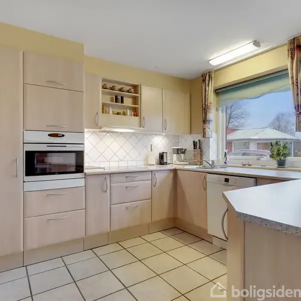 Køkken med lyse træskabe og hvide fliser, indeholdende indbygget ovn og mikrobølgeovn. Bordpladerne er dekoreret med køkkenredskaber. Gennem vinduet ses et udsigt til en have.