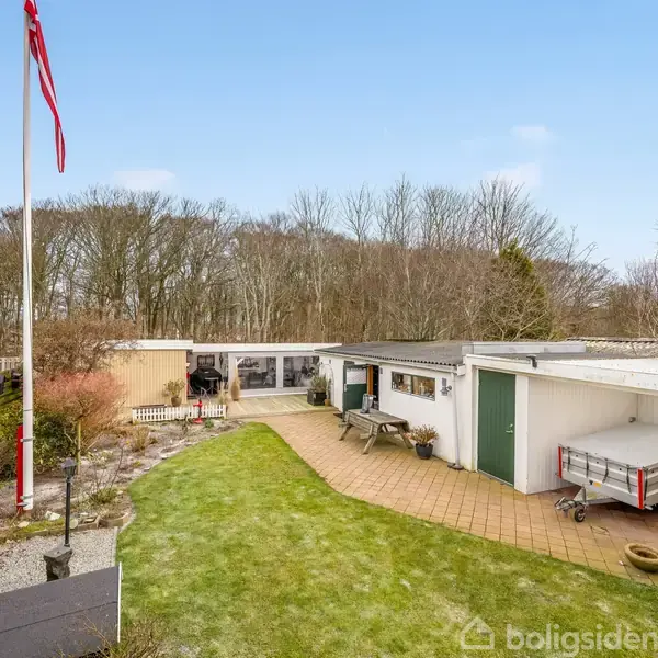 Huset står i en grøn have med stier, omgivet af træer. En trailer er placeret ved garageanlægget. Et flag blafrer i vinden ved siden af en grussti.