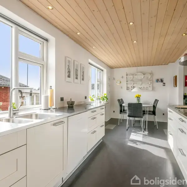 Et moderne køkken med hvide skabe og skuffer, sort gulv og træloft. En spiseplads med glasbord og sorte stole er placeret ved et vindue med udsigt til en murstensbygning.
