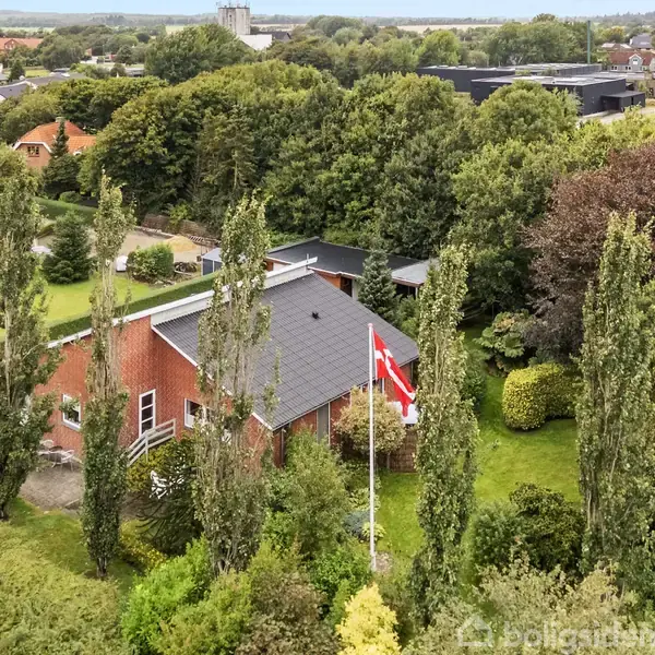 Rødt murstenshus med sort tag, omkranset af høje træer og grønne buske. En dansk flagstang står foran huset. Det bebyggede område ligger i et frodigt landskab med flere huse i baggrunden.
