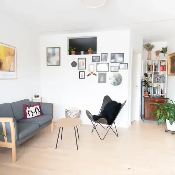 En stue med en grå sofa og en sort stol omkring et lille bord. Væggen er dekoreret med billeder. Til højre er en bogreol og planter.