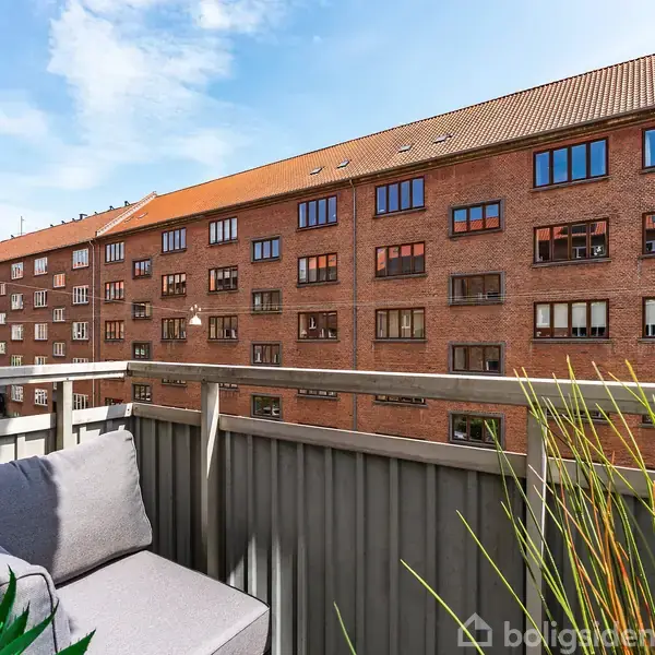 En altan med en grå hjørnesofa vendt mod et rødt murstensbyggeri i baggrunden under en blå himmel. Der er planter ved altankanten.