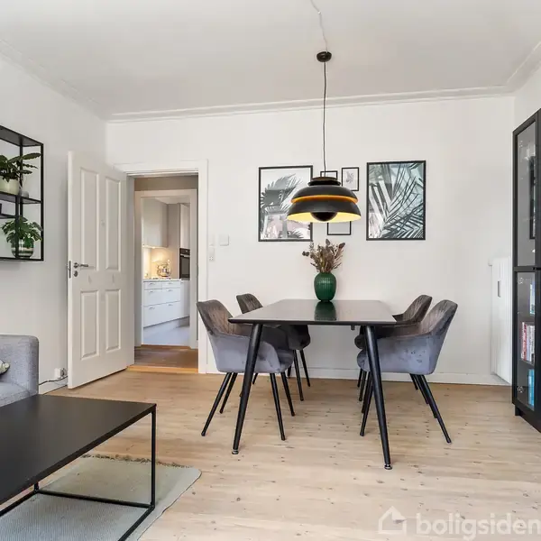 Et stilfuldt spisebord med fem stole står i en moderne stue. Omgivet af en glasbogreol, billeder på væggen og en loftslampe; dør åbner til et køkken.