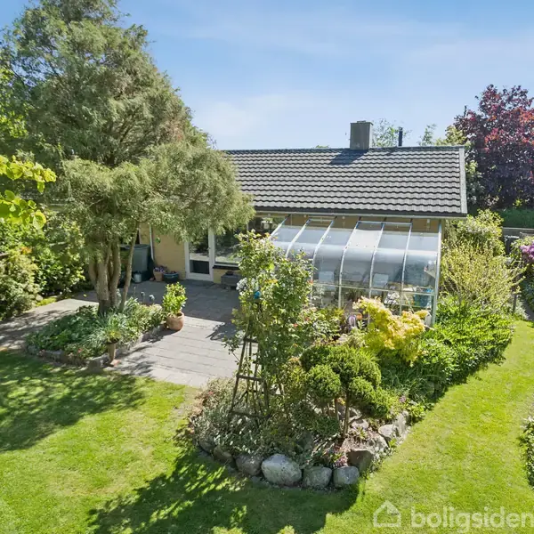 Et hus med glastilbygning ligger i en frodig have omringet af grønne træer og buske. En stenbelagt terrasse ses foran, og græsset er velplejet under en blå himmel.