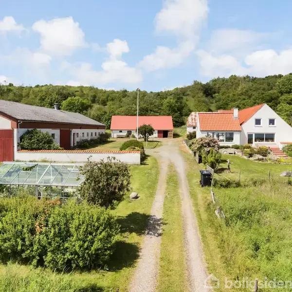 En gård med flere bygninger, herunder et drivhus, ligger langs en grusvej. Omgivet af grøn natur og skov, viser billedet en fredfyldt landlig scene.