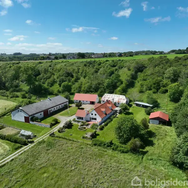Bondegård med røde tage ligger i frodigt grønt landskab. Omkringliggende bygninger omfatter ladestand med mark til venstre. Skyfri himmel og skovklædt baggrund.