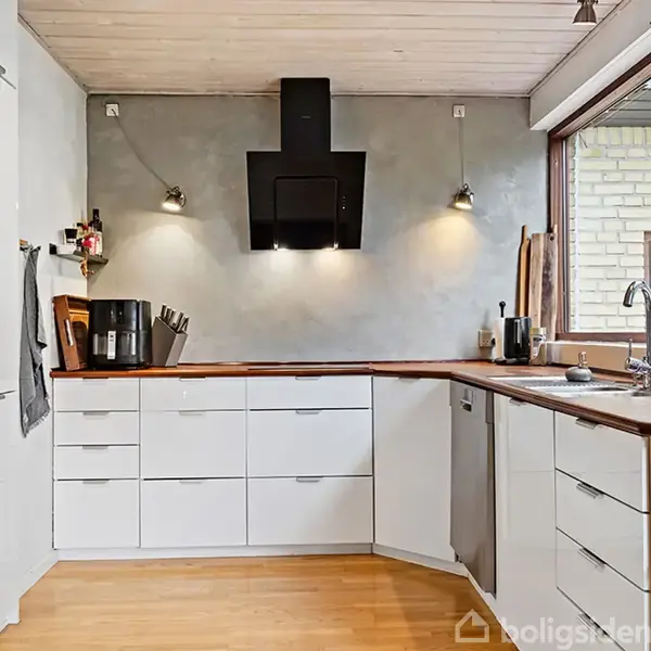 Moderne køkken med hvide skabslåger og træbordplader. En sort emhætte er vægmonteret, omringet af væglamper. Vindueskarmen har grønne planter. Ovn og kaffemaskine er synligt placeret.