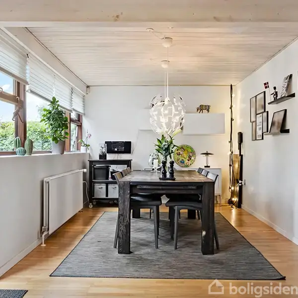 Spisebord står i et lyst rum med træmøbler og gråt tæppe, omgivet af hvide vægge. En moderne lampe hænger over bordet, og store vinduer viser grønne planter udenfor.