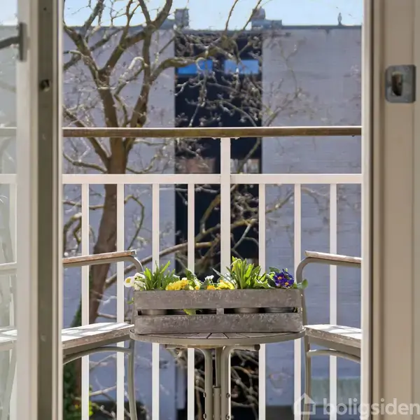 To stole omkranser et lille bord med blomsterkasse med farverige blomster. Balkonen er indrammet af en hvid gelænder, og i baggrunden ses en bygning og et træ.