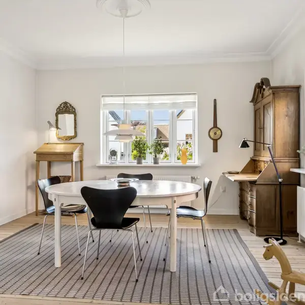 Bord med fire sorte stole står midt i et lyst rum med vinduer, der giver udsigt til en have. Et skrivebord og en kommode står mod væggen; en gyngehest står på gulvet.