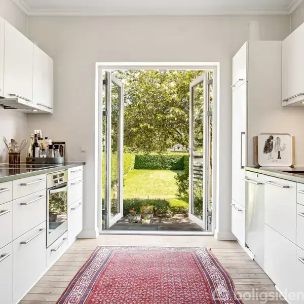 Hvidt køkken med sorte bordplader åbner mod en have gennem dobbeltdøre. En rød løber ligger på trægulvet, og der er hvide skabe på begge sider.