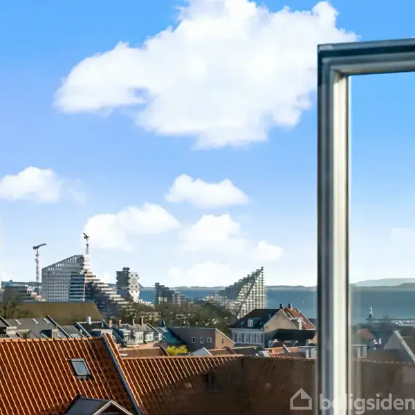 Et åbent vindue viser byens skyline med moderne og traditionelle bygninger under en blå himmel med skyer. Fjern udsigt over en blanding af ældre tage og nye højhuse.