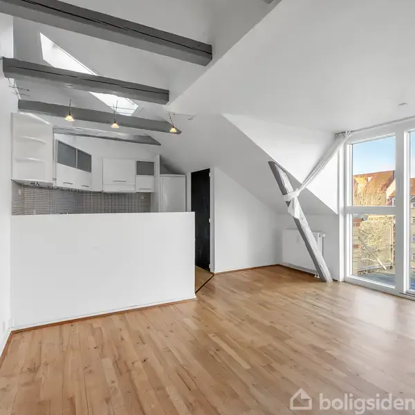 Et lyst rum med trægulv, åbent til køkken med hvide skabe. Store vinduer viser et nærliggende bygningstag. Skrå lofter med synlige bjælker tilføjer karakter.