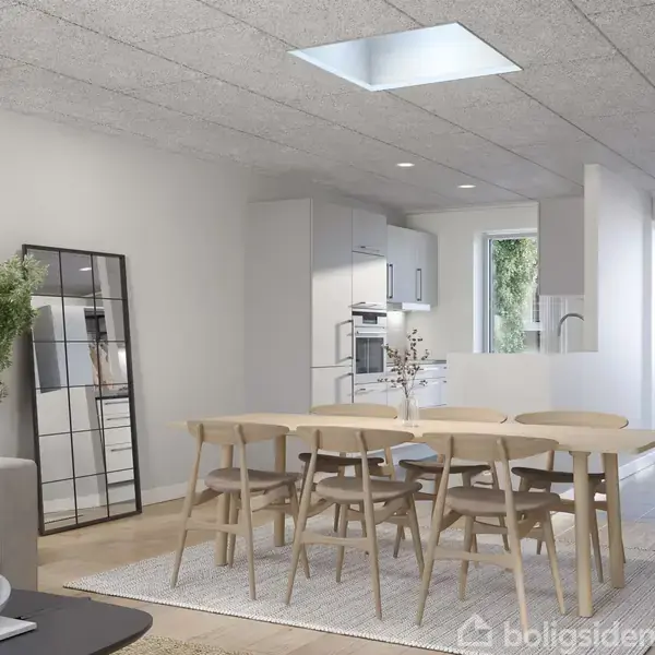 En spisestue med et lyst træbord og otte stole omgivet af en lys stue med en sofa, et spejl, og en plante. Et moderne køkken ses i baggrunden.