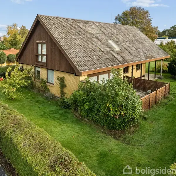 Et hus med brun træbeklædning og gul murstensfacade ligger stille i en velplejet have, omkranset af grønne buske. Der ses en indhegnet terrasse og en asfaltvej ved siden.