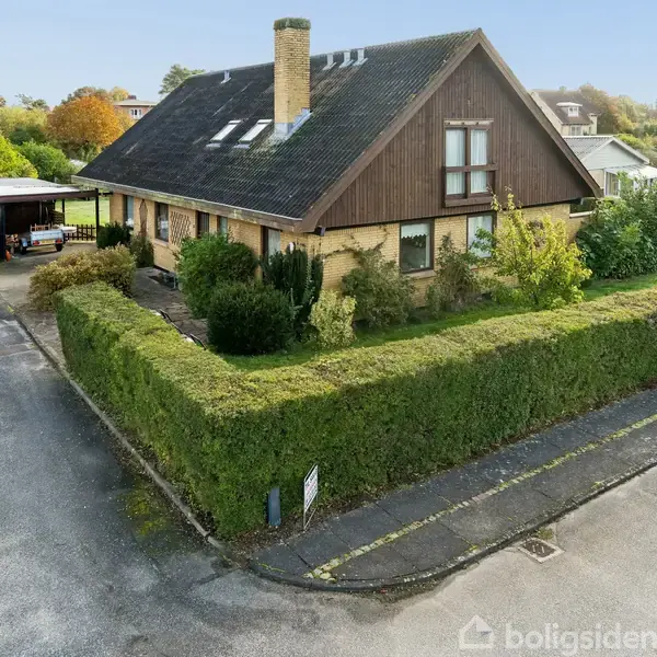 Et parcelhus med skråt tag og skorsten, omgivet af hække i et roligt boligområde. Garagen ved siden af indgangen er delvis dækket. Der er ingen tekst.