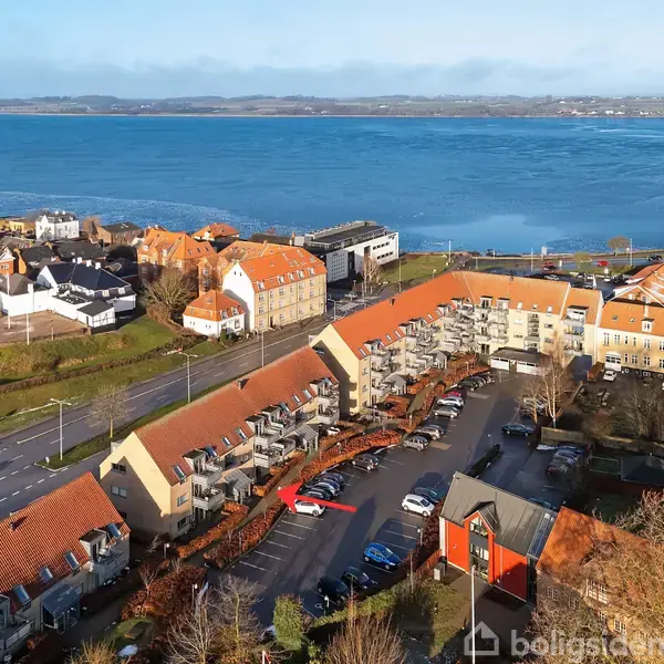 Bygninger med rødt teglstenstag står i en bue omkring en parkeringsplads. Havudsigt over blåt vand i baggrunden. Boligområde med træer og veje omkranser området.
