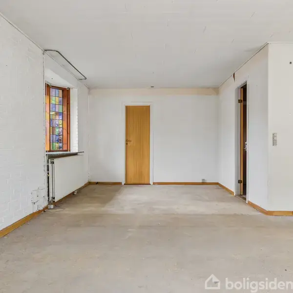 Et tomt rum med et hvidt gulvtæppe og hvide vægge. En brun dør står for enden, og et vindue med farvet glas findes til venstre, ved siden af en radiator.