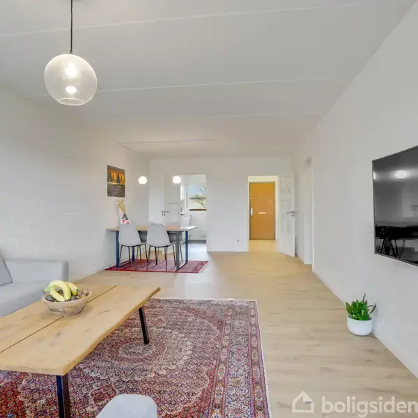 Sofaen står i en lys stue, ved siden af et træbord med skål. En fladskærm hænger på væggen. I baggrunden ses spisebord og stole på et tæppe.