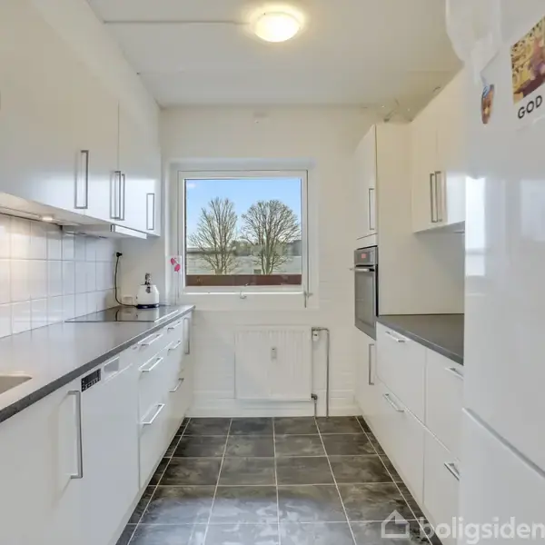 Køkken med hvide skabe langs væggene, en vask og kogeplader. Et vindue viser træer udenfor. På køleskabet hænger et kort med teksten "GOD JUL".